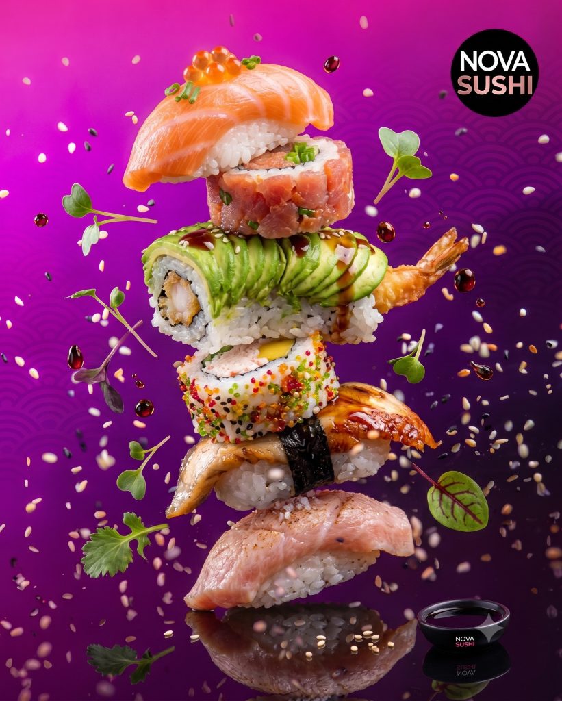 Wieża rolek i nigiri Nova Sushi w locie, na różowo-fioletowym tle, z sezamem i kroplami sosu.